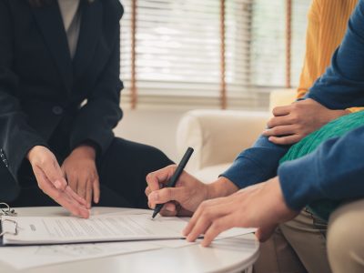Happy young Asian couple and realtor agent. Cheerful young man signing some documents while sitting at desk together with his wife. Buying new house real estate. Signing good condition contract.